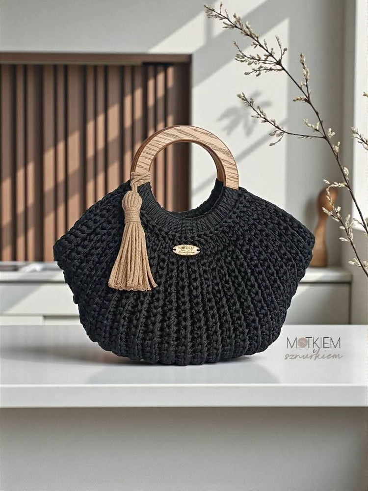 A black chunky knit handbag with a light wood top handle and a beige tassel accent, displayed on a white surface in a modern interior setting.

