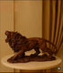 A dark brown wooden lion statue with an open mouth, displayed on a round beige pedestal against a warm beige wall and gold curtain background.

