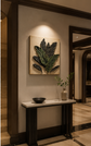A framed wall art piece featuring a 3D textured green leaf design on a beige background, displayed in a warm, modern hallway above a console table.


