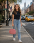 A woman walking on a city sidewalk wearing a black short-sleeve t-shirt, light-wash blue jeans, and white sneakers, carrying a pink textured handbag.

