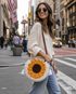 A woman wearing round sunglasses and a cream knit sweater walks on a city street, carrying a round crocheted sunflower crossbody bag with a brown strap.

